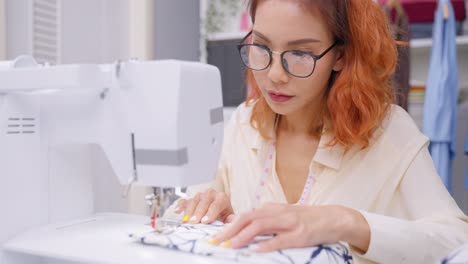 Asian-beautiful-tailor-woman-working-on-clothes-in-tailoring-atelier.-Attractive-young-female-fashion-designer-pushes-fabric-material-through-a-sewing-machine-making-new-handmade-suit-in-workshop-room