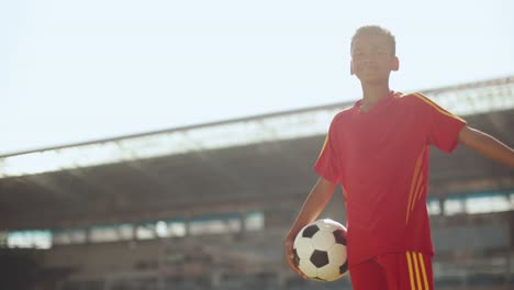 Little-boy,-child-in-red-uniform-standing-at-empty-sport-stadium-with-football-ball.-Dreaming-of-being-football-player.-Concept-of-childhood,-dreams,-imagination-and-future,-sport