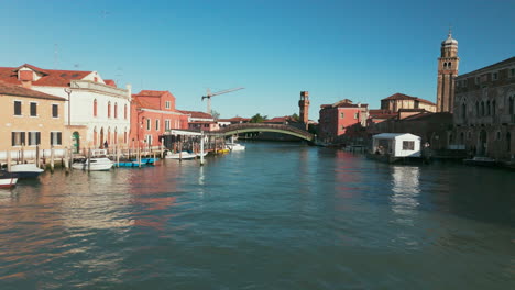 Scenic-view-of-a-canal-in-Murano,-Venice-with-a-bridge,-boats,-and-historic-buildings-on-a-sunny-day