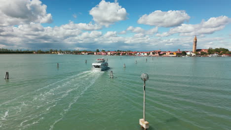 Un-Barco-De-Pasajeros-Navega-Hacia-La-Isla-De-Burano,-Italia,-Con-Sus-Edificios-Vibrantes-Y-Su-Icónica-Torre-Inclinada-Visible-En-La-Distancia.