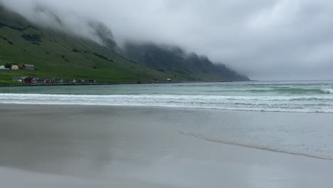 Waves-gently-lap-against-the-sandy-beach-at-Vågsøy,-Norway,-with-moody-foggy-mountains-and-colorful-wooden-houses-providing-a-picturesque-backdrop