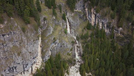 Stunning-aerial-view-of-a-waterfall-cascading-down-rocky-cliffs-in-the-Dolomite-mountains,-surrounded-by-dense-forest