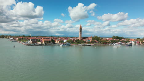 Una-Impresionante-Vista-Aérea-De-La-Isla-De-Murano,-Venecia,-Que-Muestra-El-Icónico-Campanario-Inclinado,-Barcos-Y-Edificios-Pintorescos-Bajo-Un-Cielo-Azul-Vibrante-Con-Nubes-Dispersas.