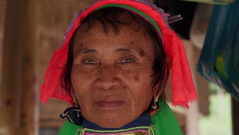 Una-Anciana-Karen-De-Cuello-Largo-Con-Anillos-De-Bronce-En-El-Cuello-Sonríe-Mientras-Posa-Bajo-Una-Choza-Al-Aire-Libre-En-Nang-Lae,-Chiang-Rai,-Tailandia,-Retrato-Con-Teleobjetivo-En-Primer-Plano