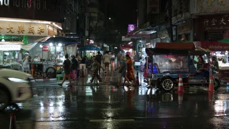 Vibrant-street-scene-of-Bangkok's-Chinatown-at-night-during-a-downpour,-with-pedestrians-and-street-vendors-along-road,-cars-drive-along-Yaowarat-Road