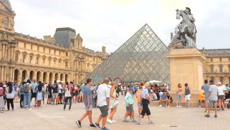 Menschenmassen-Versammeln-Sich-Vor-Dem-Louvre-Museum-In-Paris-Mit-Olympischen-Ringen-Und-Glaspyramide-An-Einem-Bewölkten-Tag