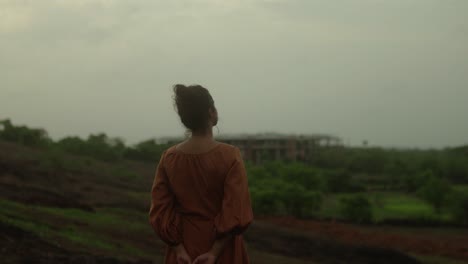 A-woman-in-a-long-dress-stands-in-a-field,-looking-at-a-distant-building-under-cloudy-skies