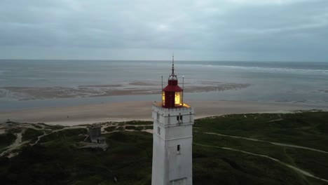 Weite-Drohnenaufnahme,-Die-Im-Uhrzeigersinn-Um-Die-Spitze-Des-Leuchtturms-Von-Blåvand-Kreist,-Mit-Eingeschaltetem-Leuchtfeuer