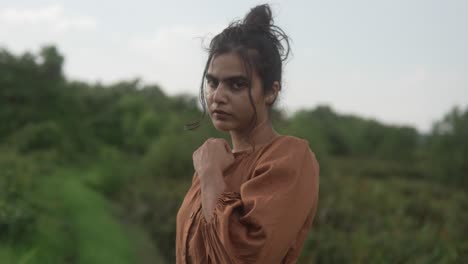 A-woman-in-a-brown-dress-stands-in-a-lush-green-field-looking-contemplative