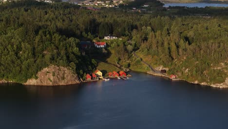 Drone-rises-above-fjord-with-homes-built-below-cliffs-as-sunset-light-casts-on-houses-by-the-dock