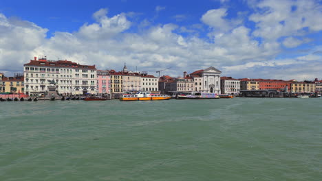 Scenic-view-of-Venice’s-waterfront-featuring-historic-buildings,-gondolas,-and-water-taxis-under-a-partly-cloudy-sky