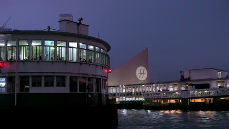 Vista-Del-Muelle-Del-Star-Ferry-Por-La-Noche-En-El-Distrito-De-Tsim-Sha-Tsui-Desde-Un-Ferry,-Con-La-Torre-Del-Reloj-De-Tsim-Sha-Tsui-Y-El-Centro-Cultural-Al-Fondo