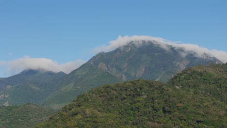 Vista-Aérea-De-Montañas-Con-Nubes-Formando-Patrones-Ondulados,-Vegetación-Densa-Y-Un-Cielo-Azul.