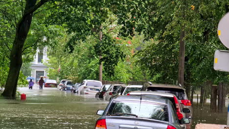 Multiple-cars-trapped-in-rising-floodwaters-on-an-urban-street,-showcasing-severe-flooding-as-friends-walk-to-observe-aftermath,-Jelgava-Latvia