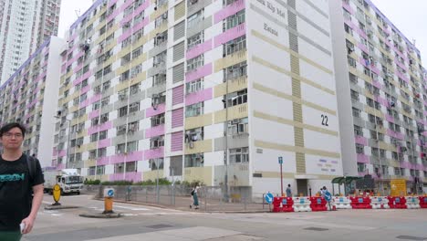 People-walk-along-the-street-as-vehicles-drive-by-with-a-colorful-high-rise-public-housing-apartment-building-in-the-background-in-Hong-Kong