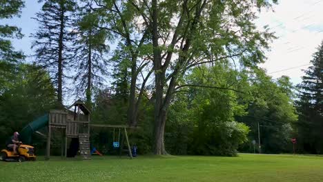 Timelapse-capturing-lawn-mowing-in-a-backyard-surrounded-by-tall-trees-and-a-children's-playset