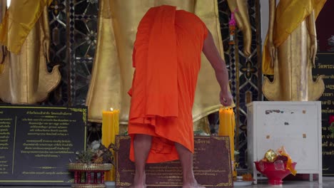 A-Buddhist-monk-in-traditional-orange-robes-lights-candles-before-a-temple-altar-at-base-of-golden-buddha-statues-and-black-plaques-at-front-of-temple