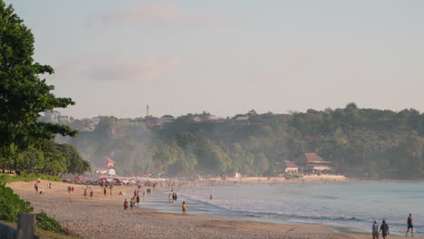 Crowded-Jimbaran-beach-with-locals-and-tourists-enjoying-the-Bali-shoreline,-Wide-angle-slow-pan-left-with-mist-in-air