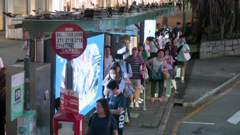 From-a-bus's-high-viewpoint,-a-nighttime-scene-in-Hong-Kong-shows-commuters-waiting-at-a-bus-stop-on-the-bustling-streets-of-Kowloon