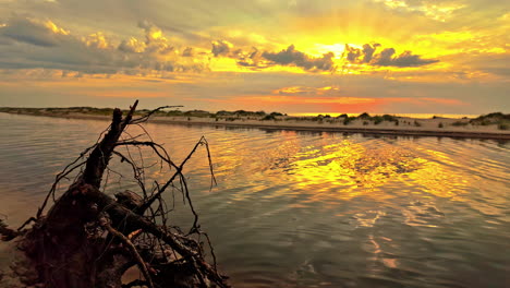 Atemberaubender-Blick-Auf-Den-Sonnenuntergang-An-Der-Küste-Lettlands-Mit-Sanddünen-Im-Wasser-Und-Einem-Erstaunlichen-Bewölkten-Himmel,-Holz-Im-Vordergrund,-Zeitlupe-Und-Kopierraum