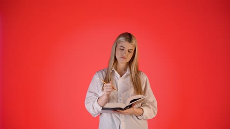 Thoughtful-Young-Blonde-Woman-Taking-Notes-On-Paper-Notebook,-Studio-Shot-With-Red-Background