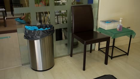 Empty-dental-clinic-waiting-room-with-metallic-bin-filled-with-medical-PPE-disposable-suits,-chair-and-small-table