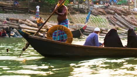 A-traditional-wooden-boat-navigates-a-river-in-Bangladesh,-with-muslim-locals-aboard-and-logs-onshore-during-sunset,-Bangladesh