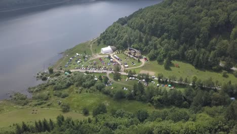 Drone-footage-of-a-car-meeting-near-a-big-lake,-surrounded-by-forest