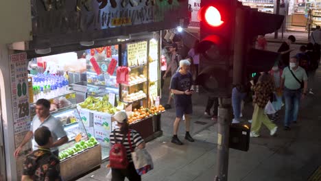 Desde-Un-Punto-De-Vista-Elevado,-Una-Escena-Callejera-Nocturna-Y-Bulliciosa-En-El-área-De-Mong-Kok,-En-Hong-Kong,-Muestra-Una-Gran-Cantidad-De-Peatones-Caminando-Frente-A-Una-Frutería,-Con-Un-Semáforo-En-Rojo-Visible-Sobre-Ellos.