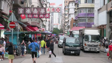 Busy-road-with-pedestrians-and-vehicles-in-the-Sham-Shui-Po-area-of-Kowloon,-Hong-Kong,-where-old-business-signs-hang-from-building-facades-above-a-street-market-and-stalls