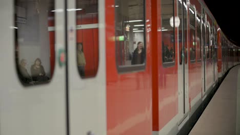 Red-train-departing-station-with-passengers-inside-and-on-platform,-evening-shot,-side-view