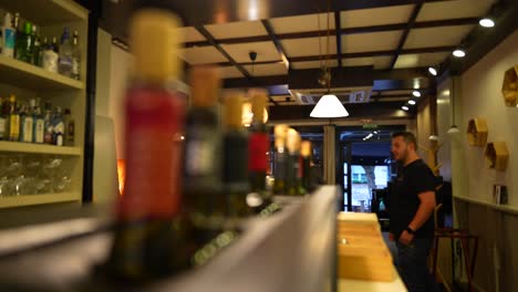 Waiter-approaches-to-cozy-bar-counter-with-warm-lighting-and-colorful-bottle-display