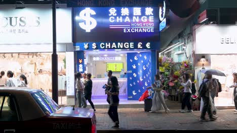 On-a-crowded-commercial-avenue-at-night,-pedestrians-pass-by-a-currency-exchange-establishment