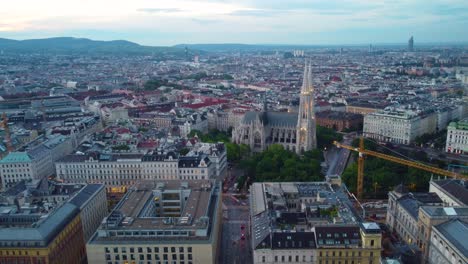 Toma-Aérea-Panorámica-Descendente-De-La-Iglesia-Votivkirche-En-Viena,-Austria,-Al-Anochecer-Con-Luces-Iluminadas-Y-Frondosos-árboles-Verdes-Debajo