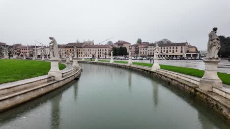 Perspektivische-Ansicht-Des-Prato-Della-Valle-Platzes-Mit-Gebäuden,-Die-Ihn-Auf-Allen-Seiten-Umgeben,-Statuen-Und-Wasser-Dazwischen-Und-Herumstreunenden-Menschen