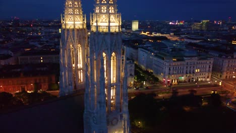 Iglesia-Votiva-Iluminada-Por-Luces-Desde-El-Interior,-órbita-Aérea-Para-Revelar-La-Ciudad-De-Viena,-Austria