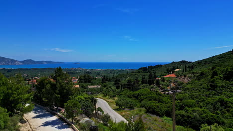 Toma-Aérea-De-Un-Pueblo-Griego-En-Una-Colina-Con-Vistas-Al-Mar,-La-Montaña-Y-Un-Horizonte-De-Verano-Despejado-Como-Fondo