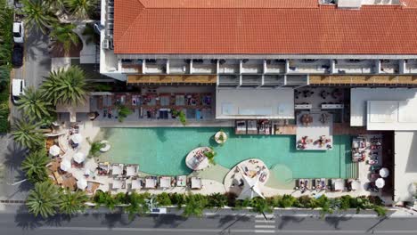 Drone-hovers-in-bird's-eye-view-over-luxury-hotel-in-Crete-Greece---A-large-pool-with-people-under-umbrellas-and-sun-loungers