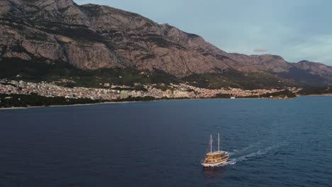 A-Boat-In-The-Adratic-Sea-With-Makarska-And-Mount-Biokovo-In-Background,-Aerial-Tracking-Shot