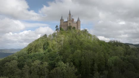 Die-Burg-Hohenzollern-Steht-Majestätisch-Auf-Einem-Waldbedeckten-Hügel-Unter-Einem-Wolkigen-Himmel,-Drohnen-Luftumlaufbahn