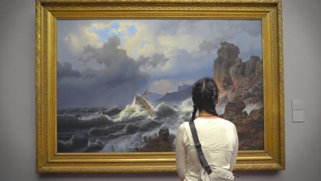 Focused-Asian-woman-observes-a-dramatic-maritime-painting-depicting-a-stormy-sea-at-the-Staedtler-Art-Museum