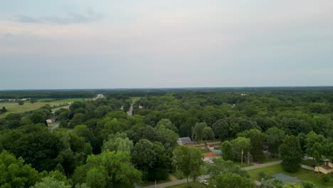 Drone-captures-a-sweeping-view-over-lush-countryside-with-scattered-buildings,-serene-greenery,-and-a-distant-lake,-offering-a-peaceful-rural-landscape