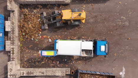 Food-waste-dumped-by-truck-and-wheel-loader-pushes-it-onto-heap,-top-down-aerial