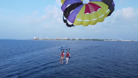 Couple-on-Summer-Vacation-Parasailing-Over-Blue-Sea,-Drone-Camera-Starts-Facing-People-and-Island-View-then-arcs-behind-to-reveal-Boat