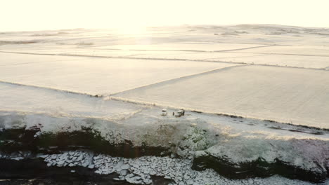 Friends-gather-at-cliff-edge-in-cars-looking-down-on-snow-covered-rocks-leading-to-ocean