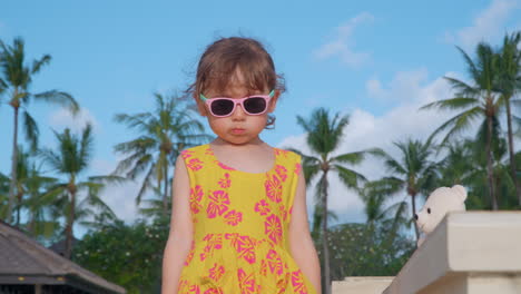 A-young-girl,-stylish-in-sunglasses-and-a-bright-yellow-dress,-enjoys-a-sunny-day-among-the-palm-trees-in-Bali-picking-up-stuffed-animal-bear-and-wiping-it-off