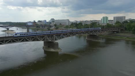 Aerial-view-of-electric-train-passing-over-bridge-on-river-in-urban-city-Warsaw