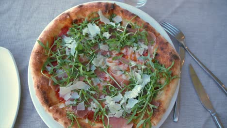 Flat-lay-shot-in-slow-motion-of-a-pizza-topped-with-cheese,-prosciutto-ham-and-rocket-salad-in-a-restaurant