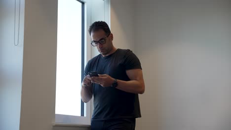 Athletic-male-in-casual-outfit-browsing-on-his-mobile-phone-while-standing-beside-an-office-window