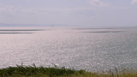 Wunderschöner-Meerblick-Von-Einer-Klippe-Mit-Der-Sonne,-Die-Sich-Auf-Der-Meeresoberfläche-Spiegelt,-Und-Langem-Grünen-Gras,-Das-In-Zeitlupe-Im-Wind-Weht,-In-Südwales,-Großbritannien,-4k